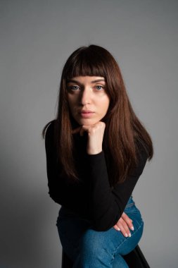 Güzel esmer bir kadının stüdyo portresi, katlanmış siyah polo yakalı bir süveter giyiyor, kameraya bakıyor, düz gri bir arka planda.