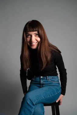 Güzel esmer bir kadının stüdyo portresini kapat, katlanmış siyah polo yakalı bir kazak giyiyor, gülüyor, yan bakıyor, düz gri bir arka plana