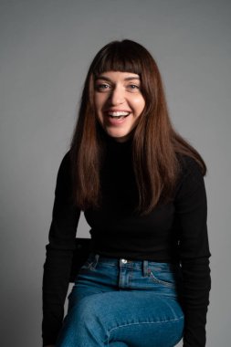 Güzel esmer bir kadının stüdyo portresini kapat, katlanmış siyah polo yaka kazak giyiyor, gülüyor, kameraya bakıyor, düz gri bir arka plana bakıyor.