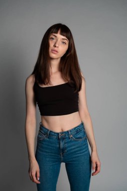 Siyah spagetti askılı güzel esmer bir kadının stüdyo portresi, kameraya ciddi ciddi bakıyor, düz gri bir arka planda duruyor.