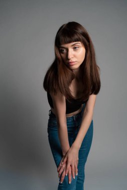 Siyah bluzlu güzel esmer bir kadının stüdyo portresi, yarı eğilmiş, iki koluyla dizine yaslanmış, düz gri bir arka plana bakıyor.