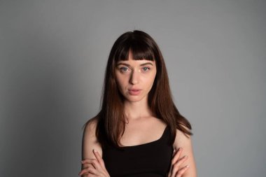 Siyah spagetti askılı güzel esmer bir kadının stüdyo portresi, kameraya ciddi ciddi bakıyor, düz gri bir arka planda duruyor.