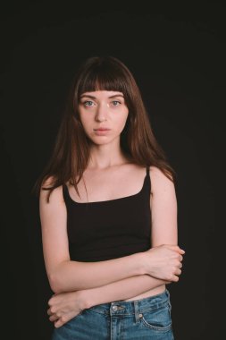 Siyah spagetti askılı güzel esmer bir kadının stüdyo portresi. Kollarını kavuşturmuş, kameraya bakıyor, düz siyah bir arkaplanda duruyor.