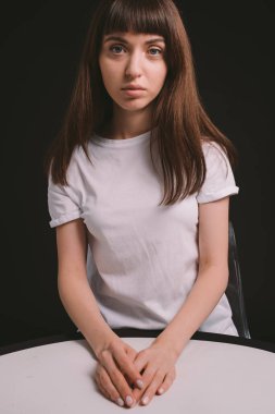 Beyaz, boş tişörtlü, siyah arka planda oturan ve kameraya bakan esmer bir kadının stüdyo portresi.