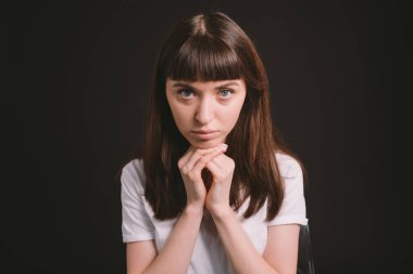 Beyaz, boş tişörtlü güzel esmer bir kadının stüdyo portresi, düz siyah bir arka plana karşı ellerini dinlendirip kameraya bakıyor.