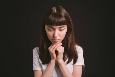 Beyaz, boş tişörtlü güzel esmer bir kadının stüdyo portresi, düz siyah bir arka plana karşı ellerini dinlendirip kameraya bakıyor.