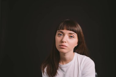 Beyaz, boş tişörtlü, siyah arka planda kameraya bakan esmer bir kadının stüdyo portresi.