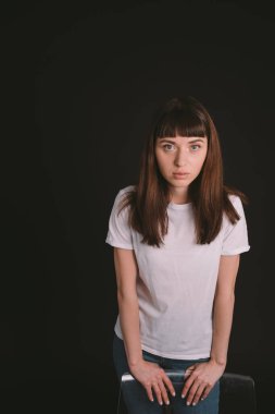 Beyaz tişörtlü güzel esmer bir kadının stüdyo portresi, bir sandalyenin arkasında, düz siyah bir arka planda, kameraya bakıyor.