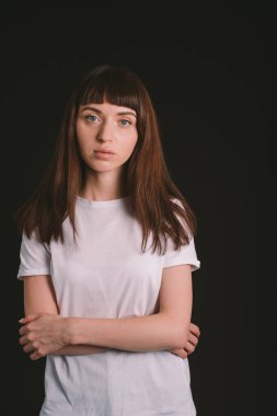 Beyaz, boş tişörtlü, kollarını kavuşturmuş, siyah bir arka planda kameraya bakan esmer bir kadının stüdyo portresi.