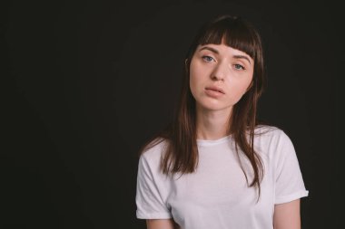 Beyaz, boş tişörtlü, siyah arka planda kameraya bakan esmer bir kadının stüdyo portresi.