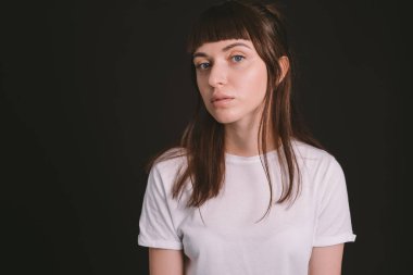 Beyaz, boş tişörtlü, siyah arka planda kameraya bakan esmer bir kadının stüdyo portresi.