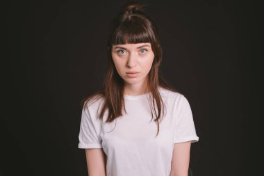 Beyaz, boş tişörtlü, siyah arka planda kameraya bakan esmer bir kadının stüdyo portresi.