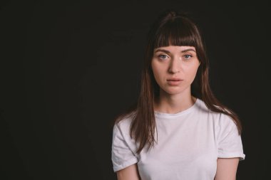 Beyaz, boş tişörtlü, siyah arka planda kameraya bakan esmer bir kadının stüdyo portresi.