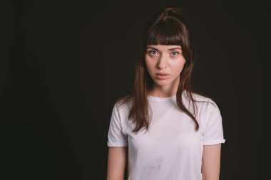 Beyaz, boş tişörtlü, siyah arka planda kameraya bakan esmer bir kadının stüdyo portresi.