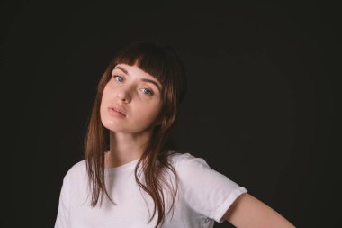 Beyaz, boş tişörtlü, siyah arka planda kameraya bakan esmer bir kadının stüdyo portresi.