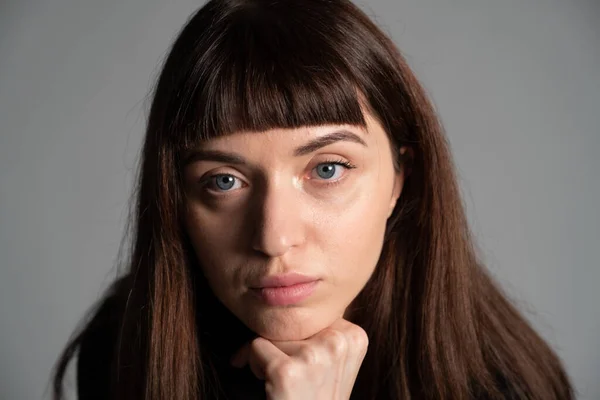 Güzel esmer bir kadının stüdyo portresini kapat, el çenesinde, ciddi ciddi kameraya bakıyor, düz gri bir arka planda.