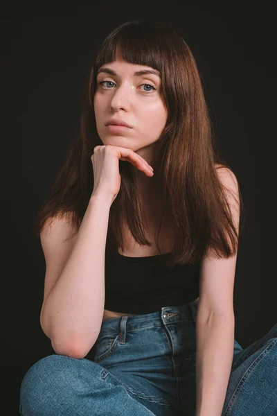 Studio portrait of a pretty brunette woman in black spaghetti strap top and jeans, looking at the camera, sitting, legs crossed, against a plain black background
