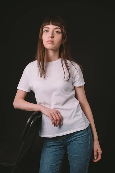 工作室的肖像一个漂亮的黑发女人在白色的空白T恤 面对着一个普通的黑色背景 看着相机 — 图库照片