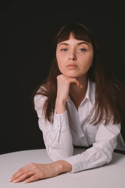 Beyaz gömlekli güzel esmer bir kadının stüdyo portresi, masada, düz siyah bir arka planda oturuyor, ciddi ciddi kameraya bakıyor.