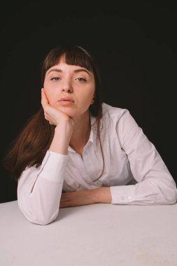 Beyaz gömlekli güzel esmer bir kadının stüdyo portresi, beyaz bir masaya yaslanmış, düz siyah bir arka plan, kameraya bakıyor.