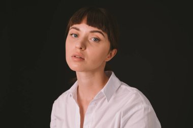 Beyaz gömlekli güzel esmer bir kadının stüdyo portresi düz siyah bir arka planda, kameraya bakıyor.