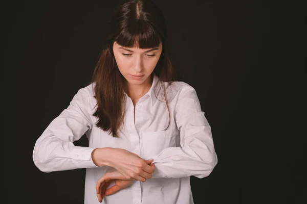 Beyaz gömlekli, siyah arka planda düğmelerini ilikleyen esmer bir kadının stüdyo portresi.