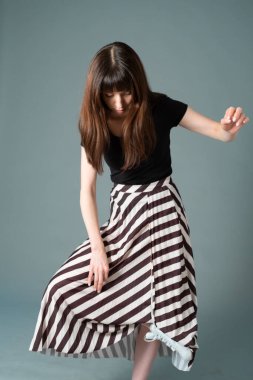 Siyah tişörtlü ve çizgili etekli güzel esmer bir kadının stüdyo portresi, tek ayak üzerinde duran, düz gri bir arka plan, aşağı bakıyor