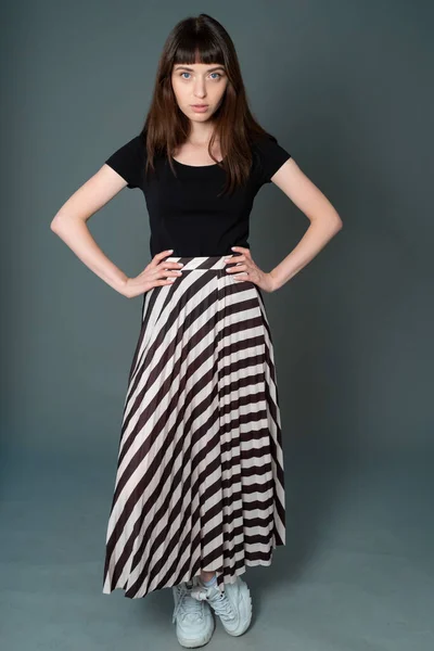 Siyah tişörtlü ve çizgili etekli güzel esmer bir kadının stüdyo portresi, düz gri bir arka planda kameraya bakıyor.