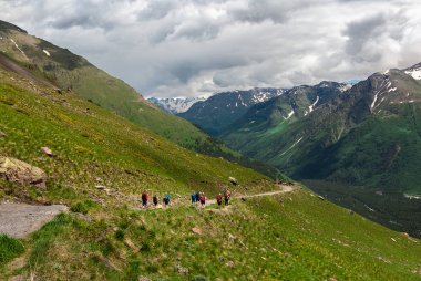 Bir grup insan, yeşil otlarla kaplı dağ lık bir dağda yürüyor. Sıcak güneşli bir gün