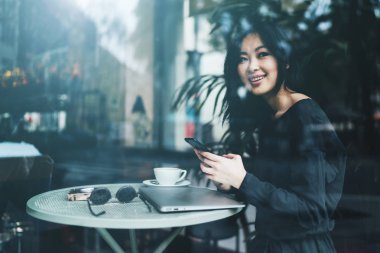 Bir cep telefonu ve dizüstü bilgisayar bir kafede otururken penceresine bakarak rahat kıyafetler giyen güzel genç kadın