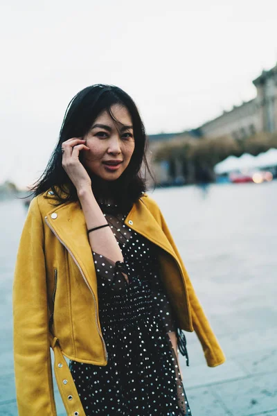 Uzun siyah saçlı bir bulanık şehir arka plan üzerinde elbise ve sarı deri ceket ayakta giyen sevimli Asyalı kadın