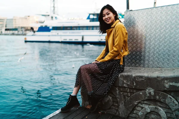 年轻迷人的亚洲女子 长着深色长发身穿雪纺裙 黄皮夹克和靴子微笑着坐在城市的泊位区域日落 — 图库照片