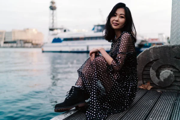 美丽迷人的亚洲妇女与长的深色头发穿着最大的雪纺礼服是微笑的相机 而坐在城市泊位区在一个模糊的游艇背景 — 图库照片
