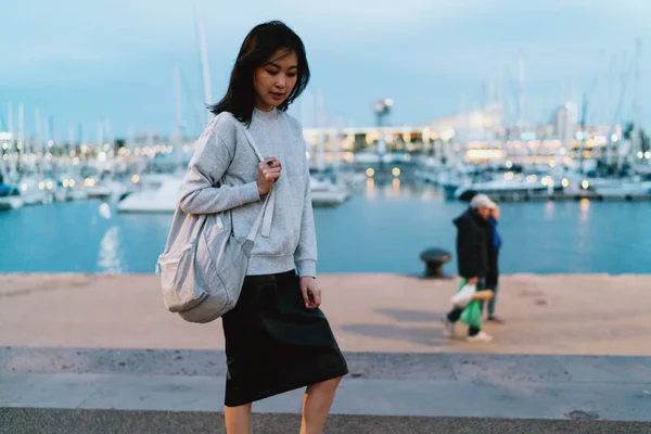 Asian woman standing with a backpack at the city berth at the evening. Toursit girl wearing gray sweatshirt visiting evening river area in the city.