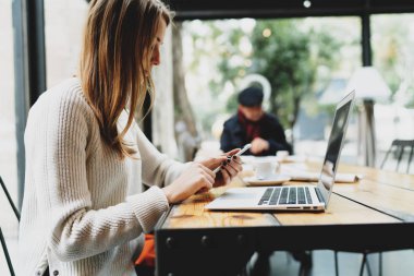 Online modern bir kafede otururken dizüstü bilgisayar ve cep telefonu kullanarak çalışan genç kadın