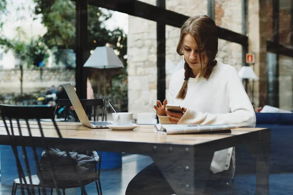 ノート パソコンや新聞のカフェに座りながら 携帯電話で同僚にメッセージングの若い女性 — ストック写真