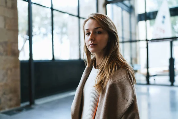 half length portrait of a beautiful blonde hipster woman looking at the camera while standing half turned to the camera on a blurred background