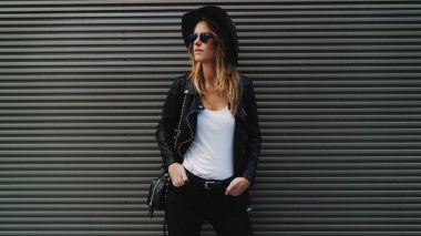 Güneş gözlüklü, uzun sarı saçlı şık hipster kız giyiyor siyah kot pantolon, deri ceket, güneş gözlüğü ve şapka bir kenara içeriğiniz için boş alanı olan gri bir duvar yanında dururken arıyorum.