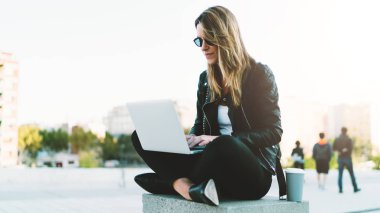Oturma ile bir parkta alırken uzak kahve bir kağıt bardak sıcak güneşli bir günde genç moda blogger giyen şık giysiler içinde yeni yazı yazma blog bir dizüstü bilgisayar tarafından wifi için bağlı..
