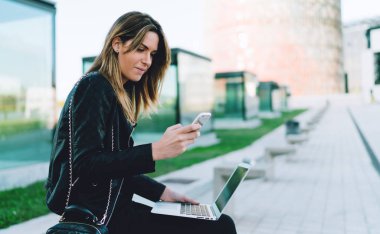 Zaman içinde bir şehir parkı güneşli bir günde tadını çıkarırken wifi için çevrimiçi bir cep telefonu ve taşınabilir bilgisayar kullanarak bilgi arama yaparak ona hafta sonu yolculuk planlama freelancer erkek bağlı
