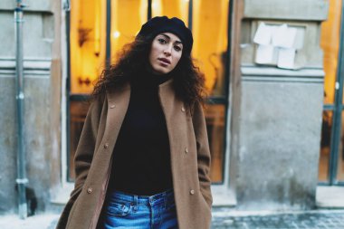 Girişimci kadın uzun siyah kıvırcık saçlı açık kahverengi palto ve akşamları sıcak ışık ile geniş bir pencere ile eski bina yanında dururken kamera bakarak siyah bere.