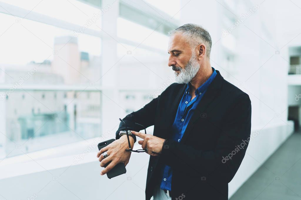 Professional estate agent checking the time on his watches while waiting for the meeting with a client in the office. Businessman waiting for the partners while standing with a mobile phone in office