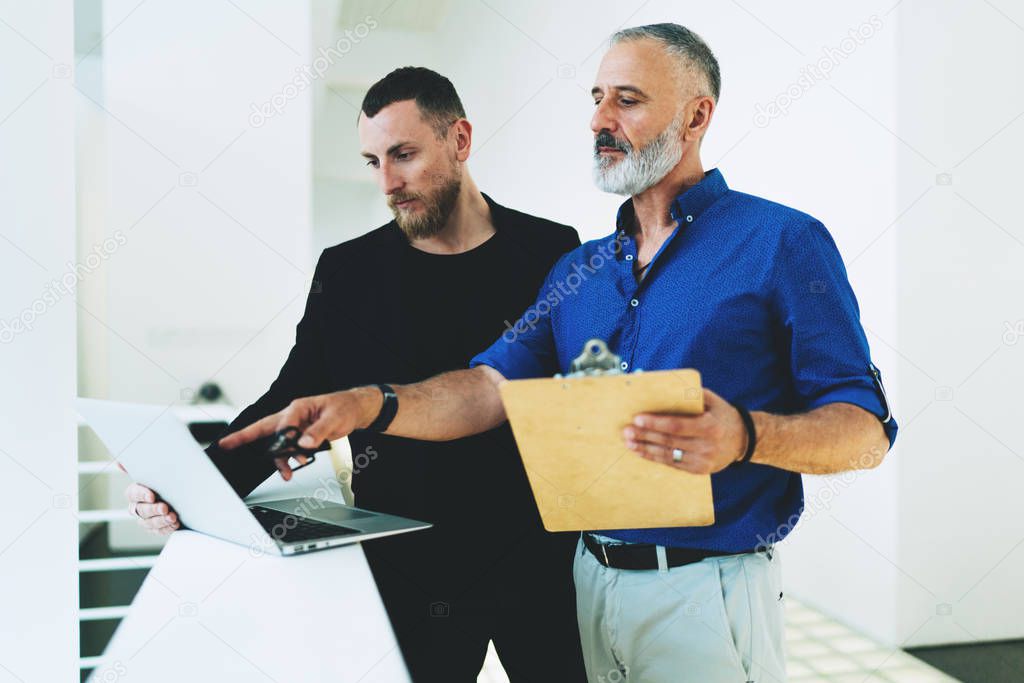 Aged male CEO discussing plan of collaboration with creative employee dressed in smart casual wear during briefing at table with laptop in stylish office. Business meeting of entrepreneurs.