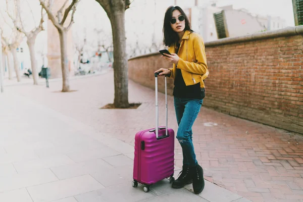 Travel blogger using internet on a mobile phone to find direction to destination. Asian hipster girl with a luggage using mobile application to call for a taxi while standing on a street.