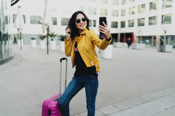 国際的な旅の間に市内中心部で自分撮り写真のポーズピンクのスーツケースを持つ魅力的なアジアの女性観光客の完全な長さの肖像画 スマートフォンのカメラで微笑む魅力的なヒップスターの女の子 — ストック写真