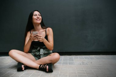 Siyah sokak duvarının yanındaki kaldırımda otururken elinde cep telefonu olan gülümseyen esmer kadın. Bir fotokopi alanının yanında poz verirken cep telefonu kullanan modaya uygun genç bir hippi kız..