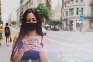 Koruyucu maske takan genç bir kadının sosyal medya üzerinden arkadaşlarıyla cep telefonuyla sohbet edişinin yarı uzunlukta görüntüsü. Siyah maskeli beyaz kadın negatif COVID19 test sonuçlarıyla e-posta aldığı için mutlu.