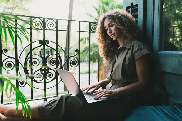 专业的博客作者在家里舒适地工作的同时 在笔记本电脑上搜索这些信息 漂亮的女人坐在一个开着的阳台旁边的地板上 膝盖上放着一本上网本 — 图库照片