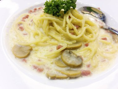 Spagetti ile hams, mantar ve peynir sosu gümüş kaşık ile beyaz tabak içinde olan maydanoz yaprakları ile dekore edilmiştir.