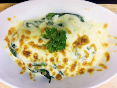 Peynirli fırında ıspanak ahşap tepsi üzerinde beyaz kase mevcuttur maydanoz yaprakları ile süslüdür.